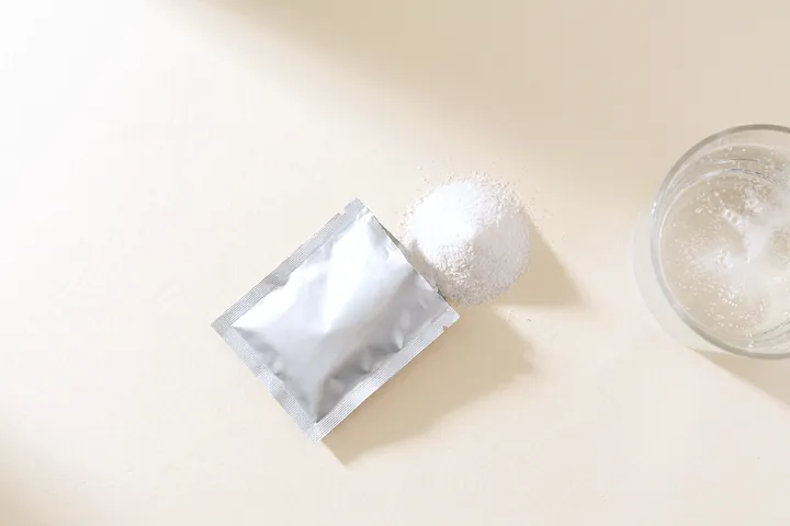 An unmarked foil sachet with white effervescent powder spilled beside it, and a glass of fizzing water